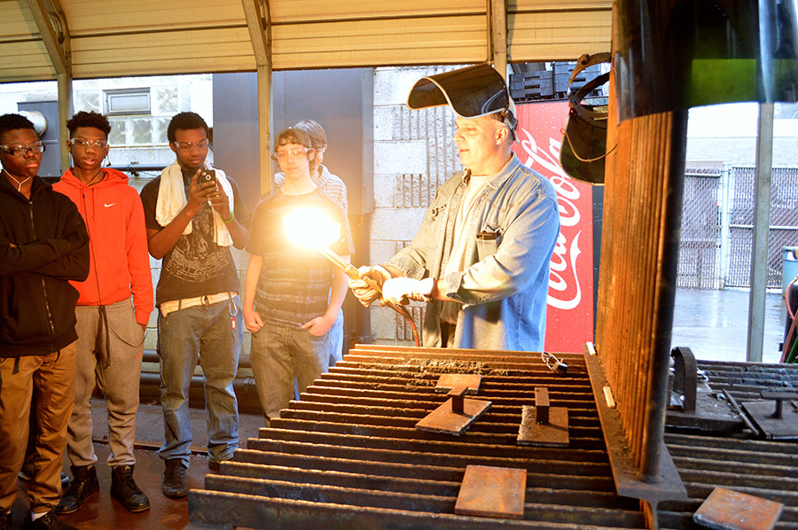 Students stand in a circle around a tradesman with a lit blowtorch